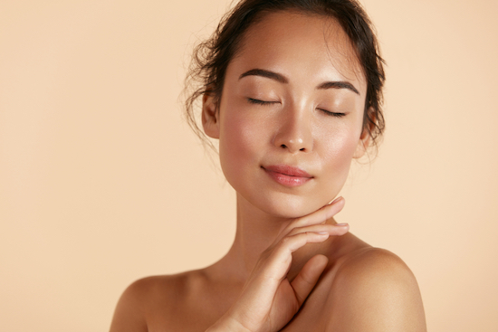 Beauty face. Woman with natural makeup and healthy skin portrait. Beautiful asian girl model touching fresh glowing hydrated facial skin on beige background closeup. Skin care concept