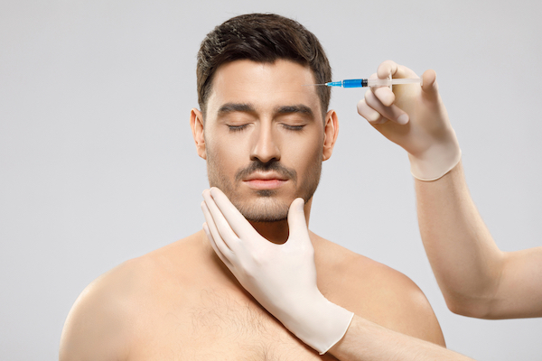 Young shirtless man receiving beauty injection in forehead to remove wrinkles, face held by hands of cosmetologist, isolated on gray background. Plastic surgery concept