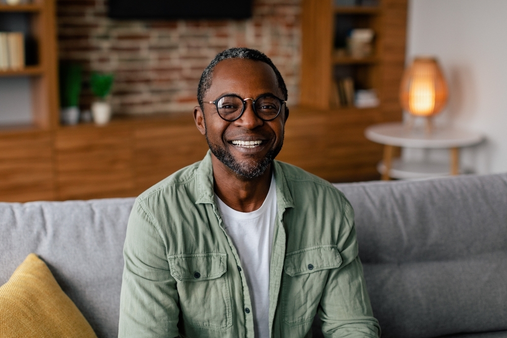Headshot of cheerful adult african american man in glasses and casual relaxing on sofa and looking at camera in living room interior, close up.