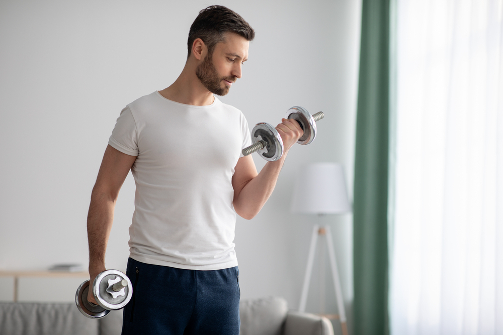 A man in a white t-shirt lifts dumbbells at home, focusing on his workout in a bright living room with a cozy atmosphere