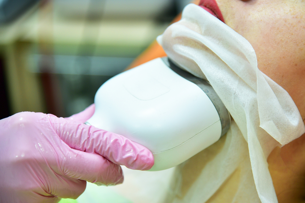 A technician in pink gloves applies a facial treatment device to a client's chin, covered with a protective cloth