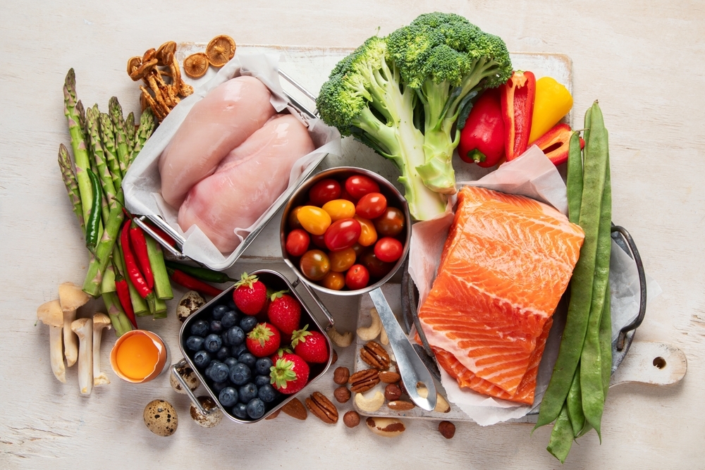 A vibrant assortment of fresh foods including chicken breasts, salmon fillet, vegetables, fruits, nuts, and spices arranged on a wooden board