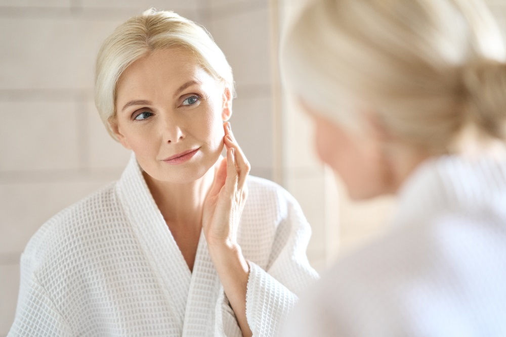 A woman in a white robe examines her skin in a bathroom mirror, illuminated by soft natural light